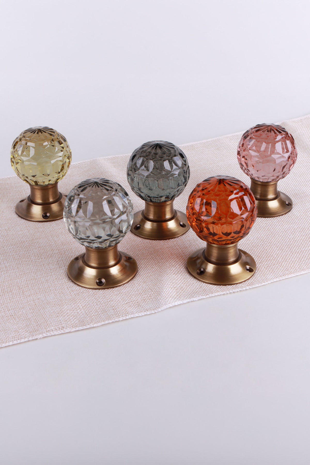 Five faceted glass door knobs on antique brass roses in ivory, clear crystal, smokey grey, amber and blush pink on a linen runner