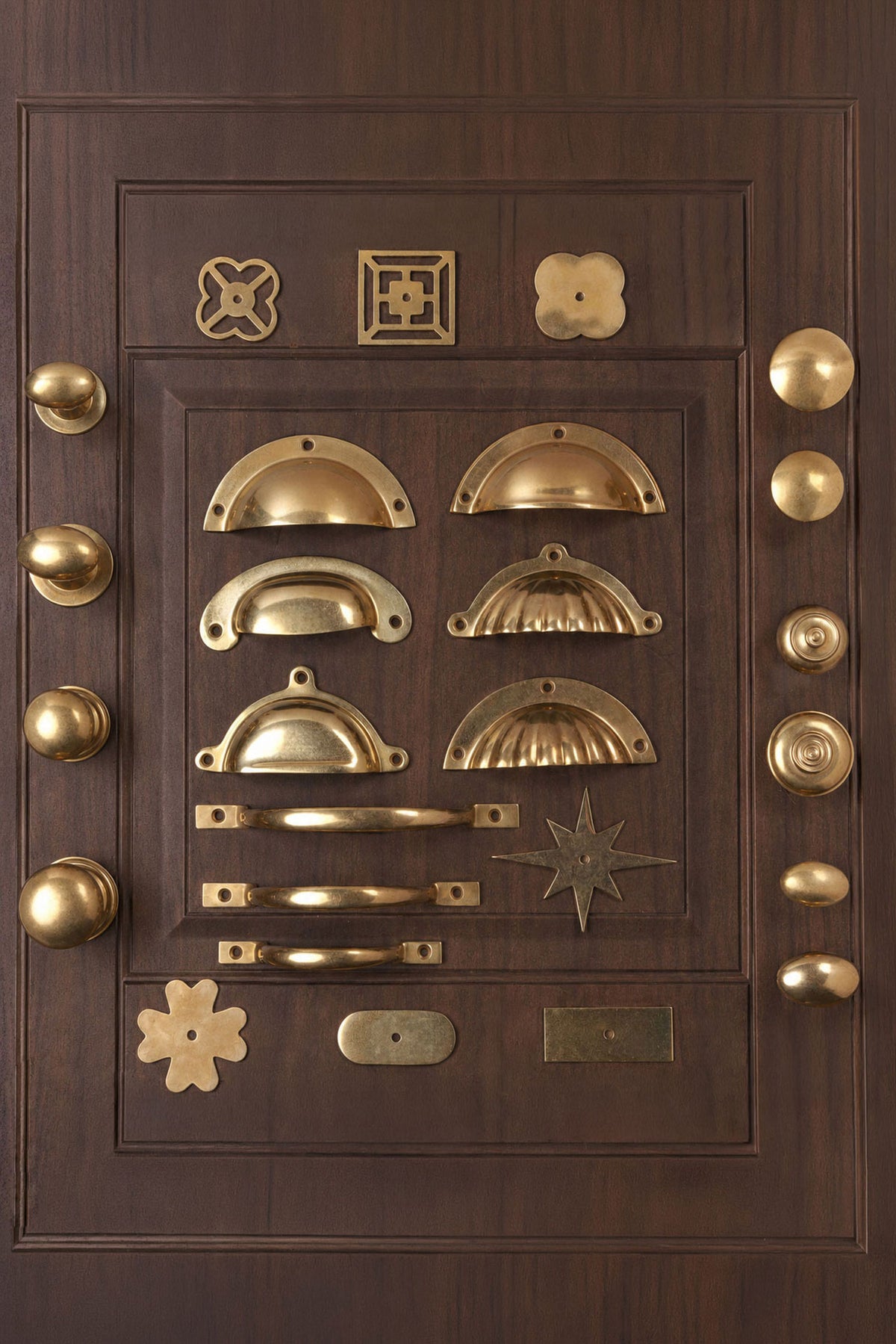 Aged brass cabinet knobs, cup pulls, D handles and backplates laid out on a panelled cupboard door