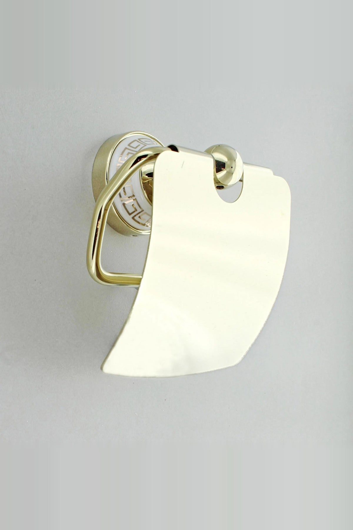 Gold toilet roll holder with a curved cover, seen from the side, on a white Greek key wall plate