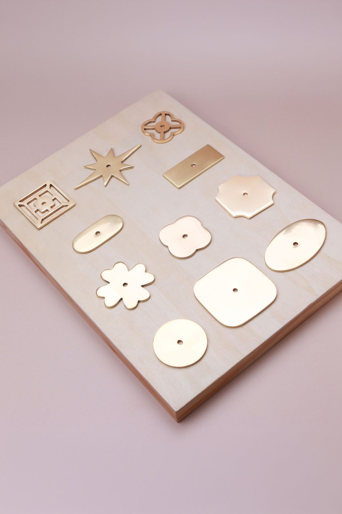 Eleven polished brass cabinet knob backplates on a pale wooden board: round, oval, square, squircle, quatrefoil, pierced, fretwork, flower, star, oblong and rectangle, second view