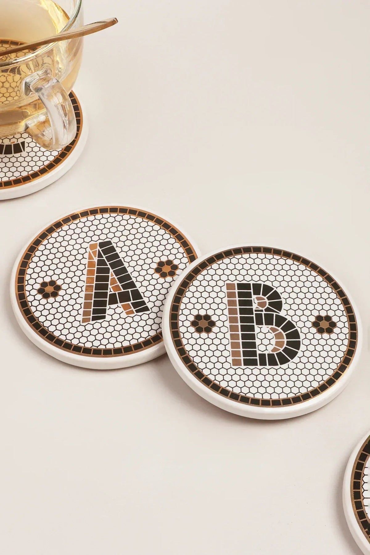 A considered table — black and gold British pub tile monogram coasters in hand-finished ceramic, a tableware detail set among glass and linen | G Decor
