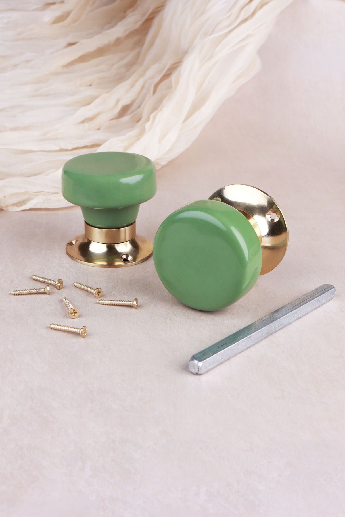 Sage green ceramic door knob standing on its polished brass rose beside the second knob, with the spindle and fixing screws