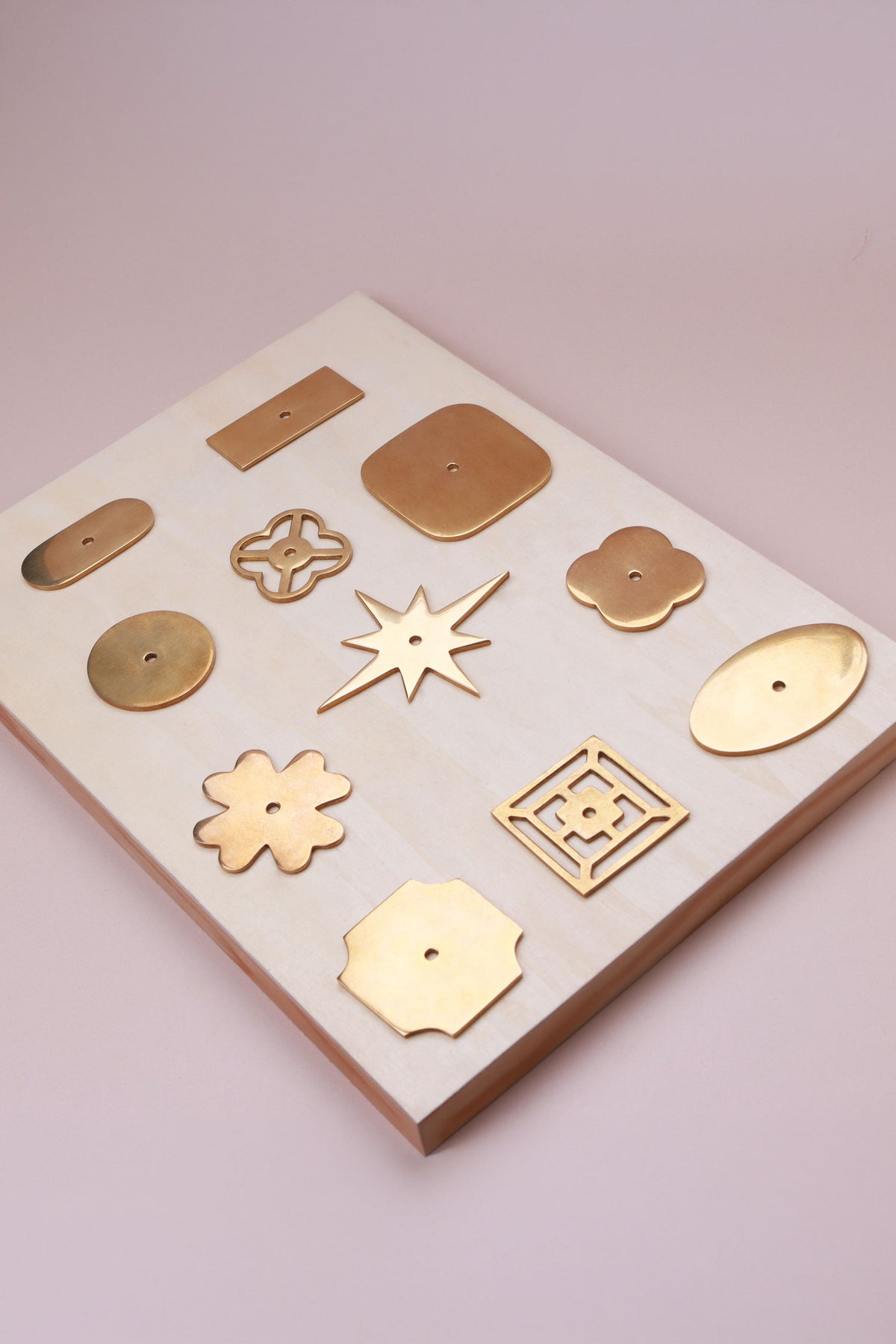 Eleven aged brass cabinet knob backplates on a pale wooden board: round, oval, square, squircle, quatrefoil, pierced, fretwork, flower, star, oblong and rectangle