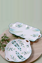Two ceramic serving plates with green and blue leaf sprays and cut-out handles, a long platter and a round plate, on a wooden board with leaves