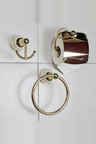 Gold bathroom set on a white tiled wall: double robe hook, toilet roll holder with cover holding a roll, and a towel ring, each on a white Greek key wall plate