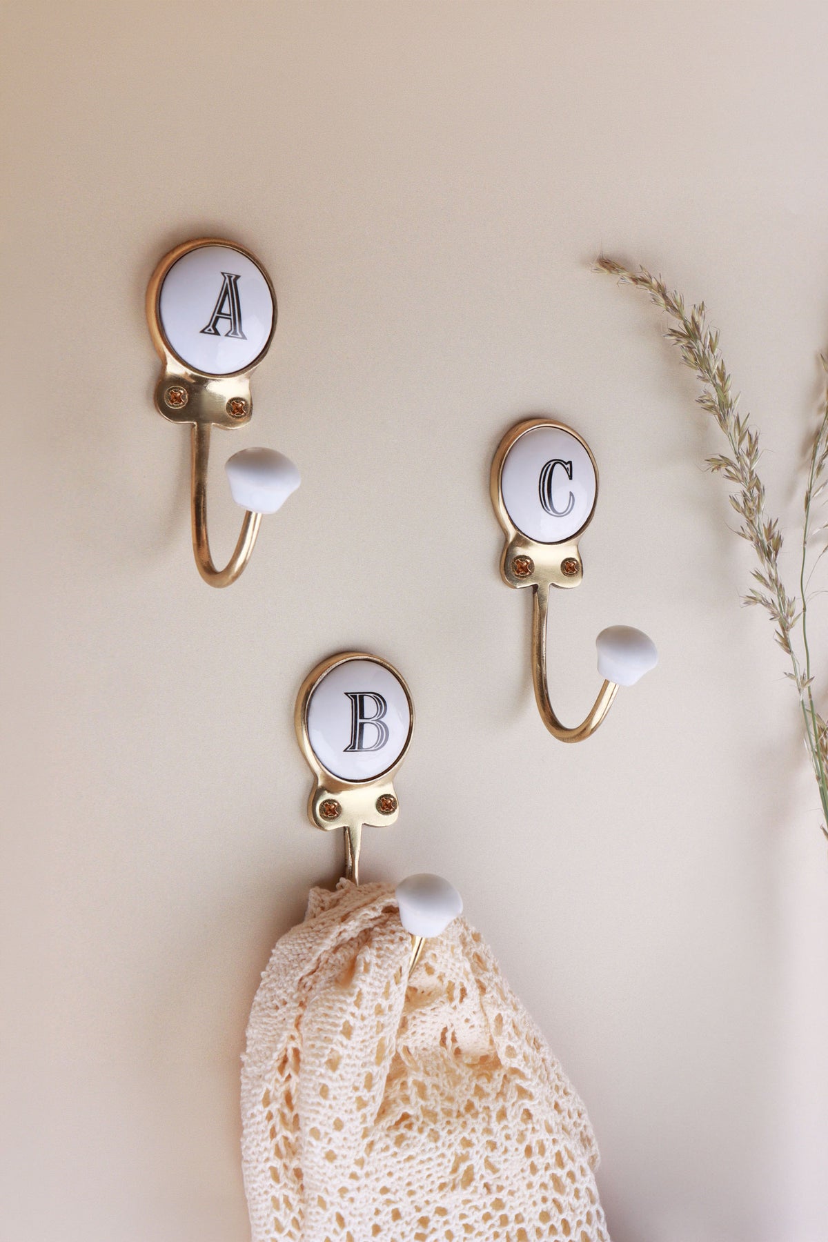 Ceramic letter hooks A, B and C on a cream wall with dried grass: white discs with black serif capitals in gold-tone frames, a lace cloth hanging from B